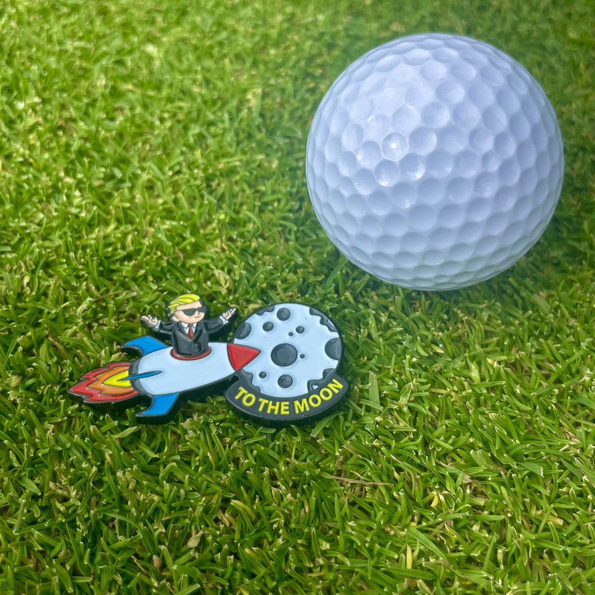 Ballmarker To The Moon von Alpha Putts neben einem Golfball auf dem Grün