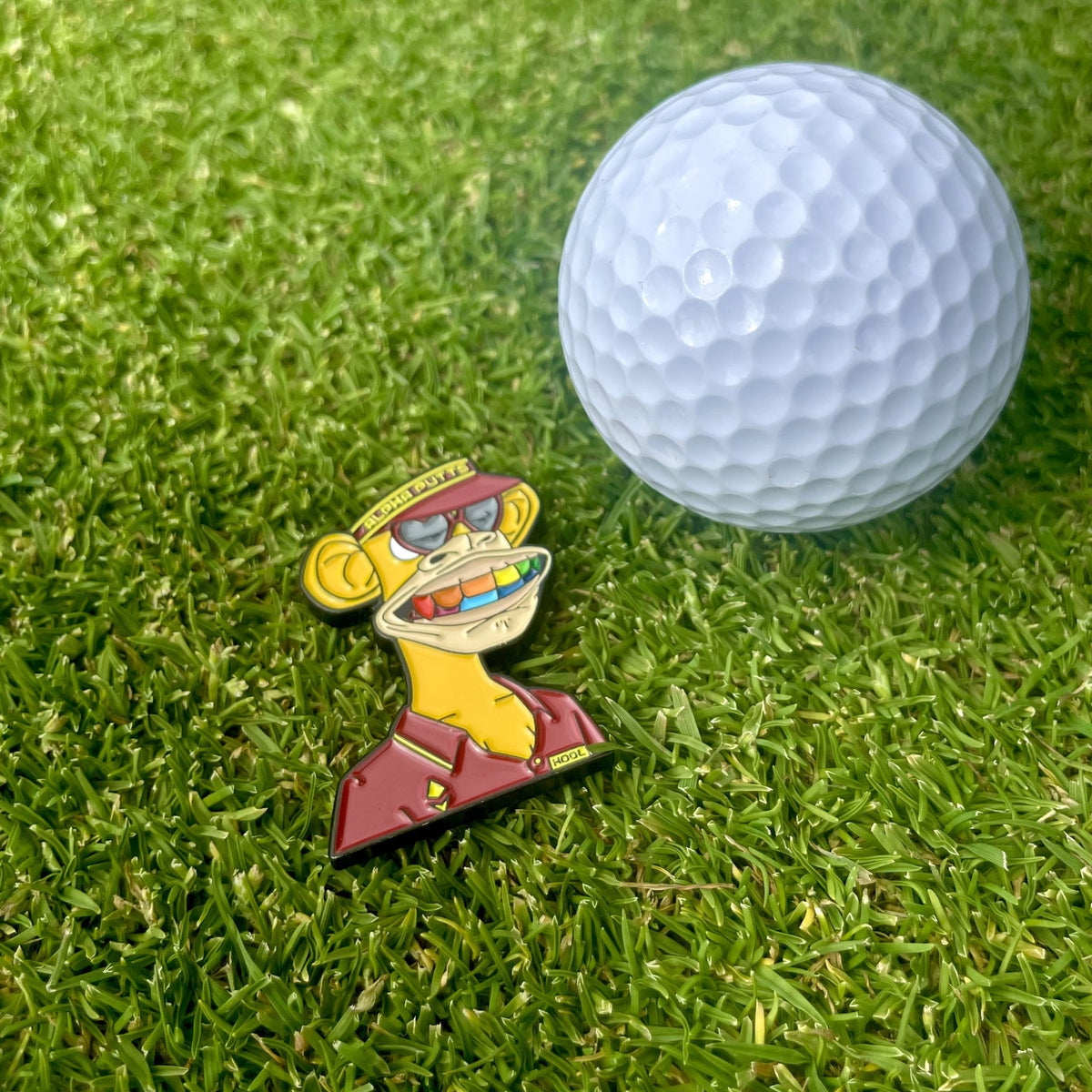 Ballmarker Bored Ape #7090 von Alpha Putts neben einem Golfball auf dem Grün